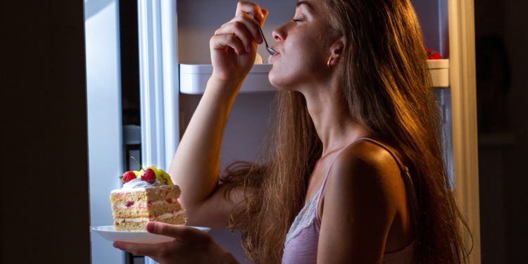 Eine Frau steht am Kühlschrank und isst ein Stück Kuchen.