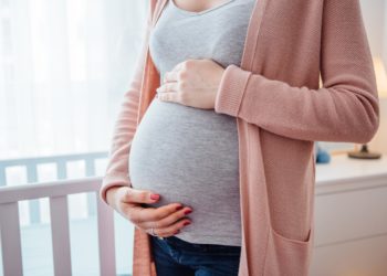 Eine schwangere Frau steht neben einem Babybett und hält mit den Händen ihren Bauch.