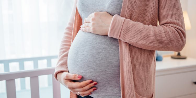 Eine schwangere Frau steht neben einem Babybett und hält mit den Händen ihren Bauch.