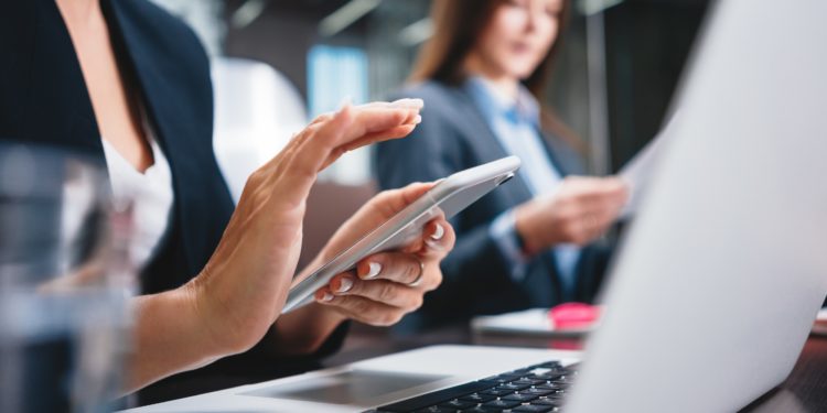 Eine Frau sitzt vor einem Laptop und schaut auf ihr Handy.