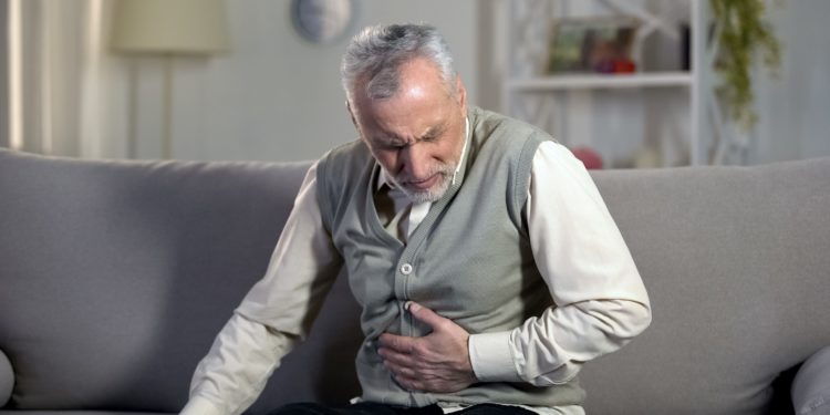 Ein älterer Mann mit schmerzverzerrtem Gesicht sitzt auf dem Sofa und hält sich die Hand an den Bauch. 