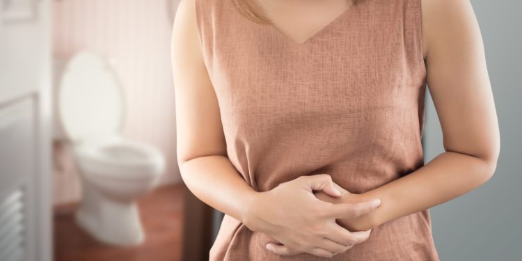 Eine Frau steht vor der geöffneten Tür zur Toilette und hält sich die Hände an den schmerzenden Bauch.