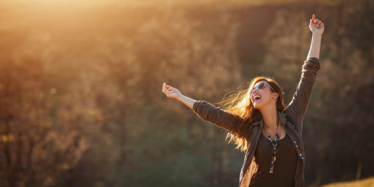 Junge Frau in einer Naturlandschaft lächelt und streckt ihre Arme in die Höhe