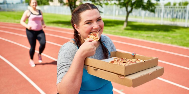 Zwei Frauen joggen über eine Laufstrecke und essen dabei Pizza.