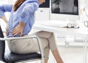 Am Schreibtisch sitzende Frau fasst sich mit ihren Händen an den schmerzenden Rücken