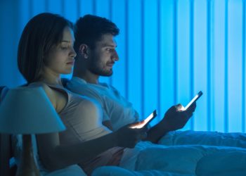 Frau und Mann sitzen nebeneinander im Bett und schauen auf ihre Smartphones