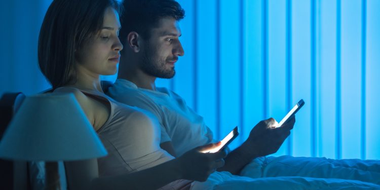 Frau und Mann sitzen nebeneinander im Bett und schauen auf ihre Smartphones