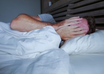 Im Bett liegender junger Mann bedeckt mit seinen Händen sein Gesicht