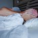 Im Bett liegender junger Mann bedeckt mit seinen Händen sein Gesicht