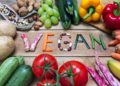 Obst, Gemüse, Nüsse und Hülsenfrüchte um den Schriftzug Vegan auf einem Holztisch