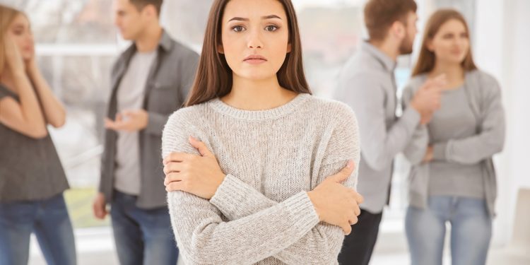 Eine Frau steht mit verschränkten Armen und ängstlichem Blick vor einer Gruppe von Männern und Frauen. 