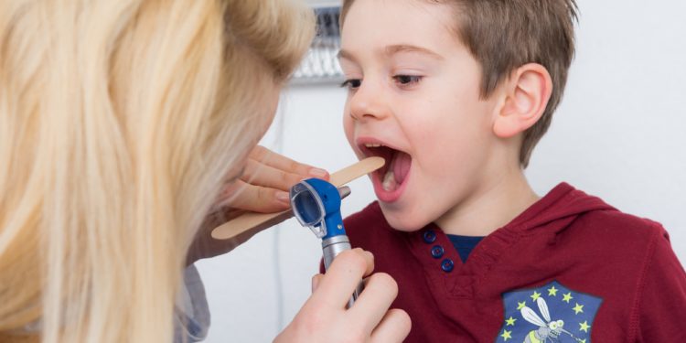 Eine Kinderärztin untersucht mithilfe einer Lampe und eines Holzspatels den Rachen eines Jungen. 