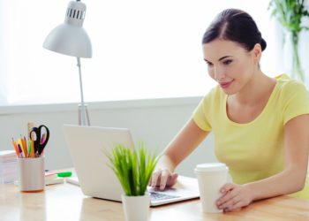 Lächelnde junge Frau sitzt mit einem Kaffee und einer Grünpflanze am Schreibtisch an einem Laptop