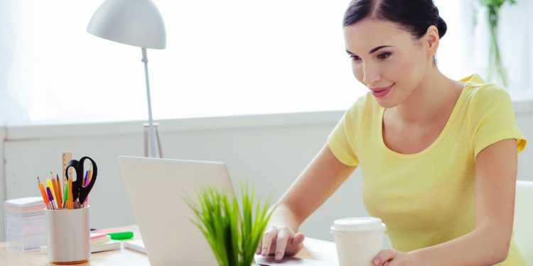 Lächelnde junge Frau sitzt mit einem Kaffee und einer Grünpflanze am Schreibtisch an einem Laptop