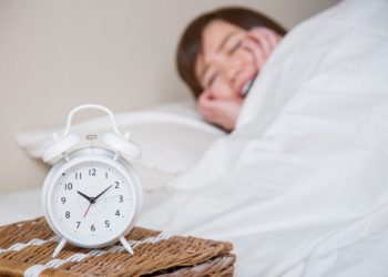 Frau liegt im Bett und hält sich die Ohren zu im Vordergrund steht ein Wecker auf zehn Minuten nach Zehn