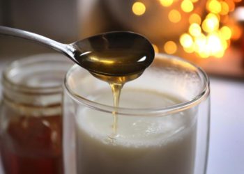 Honig fließt von einem Löffel in ein Glas mit heisser Milch.