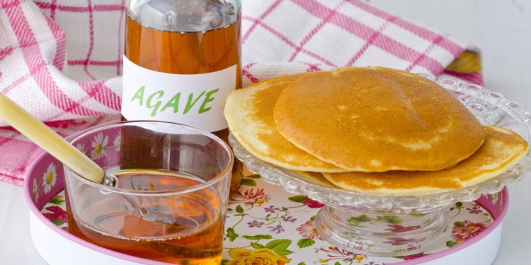 Auf einem Tablett stehen eine Flasche und ein Glasschälchen mit Agavensirup sowie eine kleine gläserne Kuchenplatte mit mehreren Pancakes.