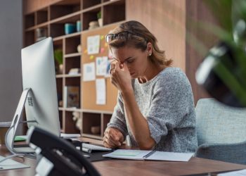 Frau im Büro hat Kopfschmerzen und fasst sich an die Nasenwurzel