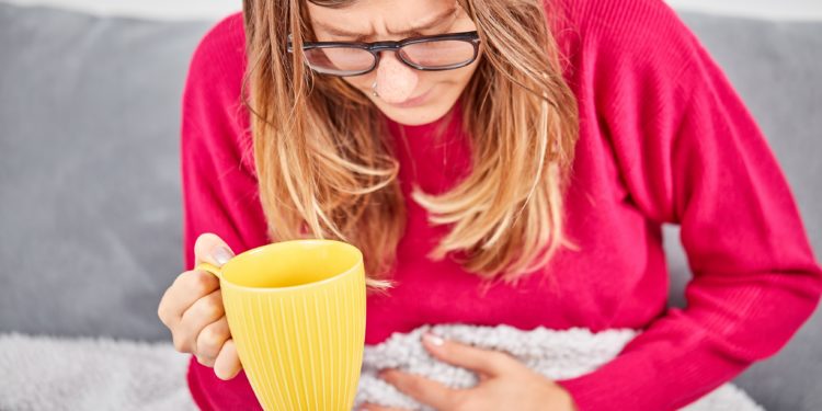 Eine Frau sitzt mit gesenktem Kopf auf dem Sofa und hält in einer Hand einen Becher, während sie mit der anderen Hand an ihren Bauch fasst.