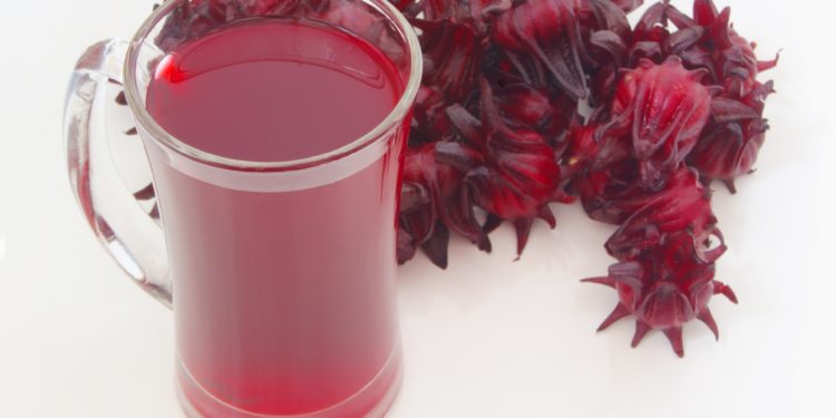 Ein Glas mit Hibiskustee und mehrere getrocknete Hibiskusblüten auf weißem Untergrund.
