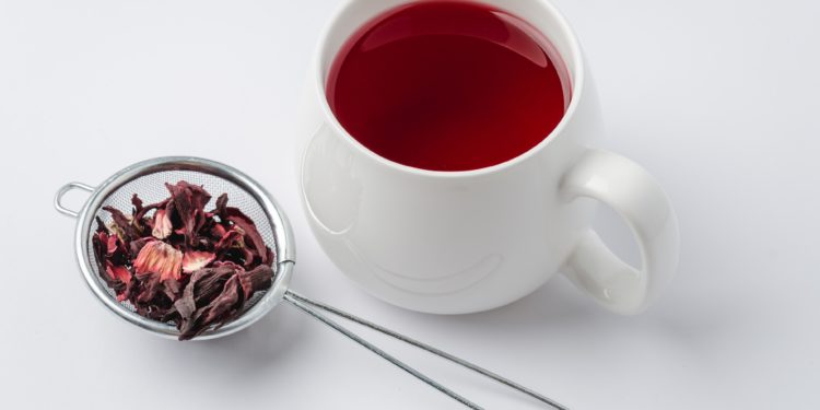 Hibiskusblüten in einem Teesieb neben einem weißen Becher mit Hibiskustee. 