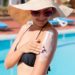 Eine Frau mit Bikini, Sonnenhut und Sonnenbrille steht an einem Swimmingpool und cremt sich den Arm mit Sonnencreme ein.