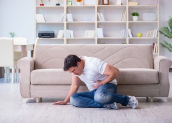 Ein Mann sitzt zusammengesunken auf dem Fußboden vor einem beigen Sofa und hält sich die Hand an den Bauch.