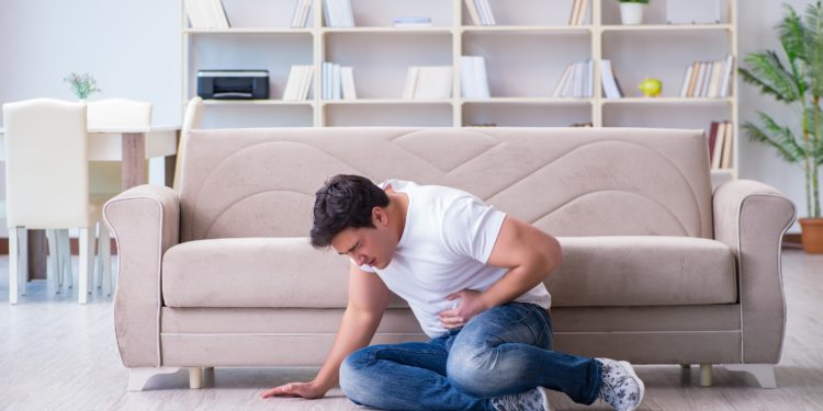 Ein Mann sitzt zusammengesunken auf dem Fußboden vor einem beigen Sofa und hält sich die Hand an den Bauch.