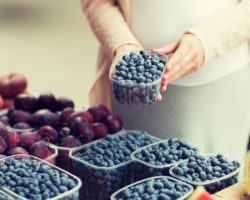 Eine schwangere Frau steht an einem Obststand und hält eine Plastikschale mit Heidelbeeren in den Händen.