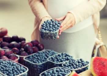 Eine schwangere Frau steht an einem Obststand und hält eine Plastikschale mit Heidelbeeren in den Händen.