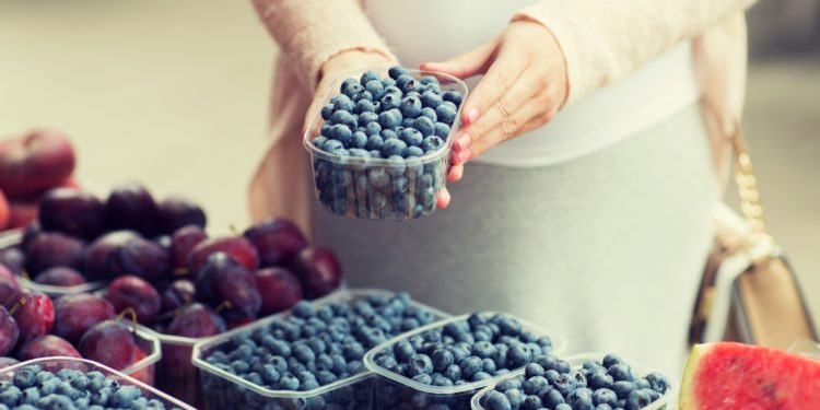 Eine schwangere Frau steht an einem Obststand und hält eine Plastikschale mit Heidelbeeren in den Händen.