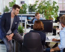 In einem Großraumbüro sitzen mehrere Mitarbeiterinnen und Mitarbeiter an einem Tisch.