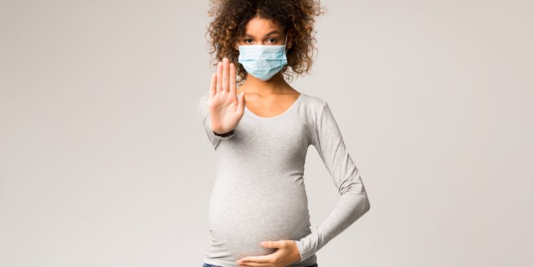 Eine Schwangere trägt eine Atemschutzmaske und macht eine abwehrende Geste.