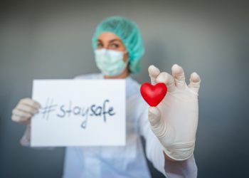 Eine Ärztin hält ein Plastikherz in der einen Hand und in der anderen Hand ein Papier mit der Aufschrift "stay safe".