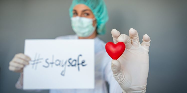 Eine Ärztin hält ein Plastikherz in der einen Hand und in der anderen Hand ein Papier mit der Aufschrift "stay safe".