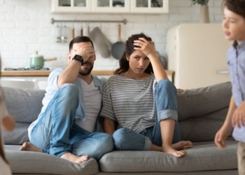 Genervte junge Eltern auf dem Sofa und zwei herumtollende Kinder