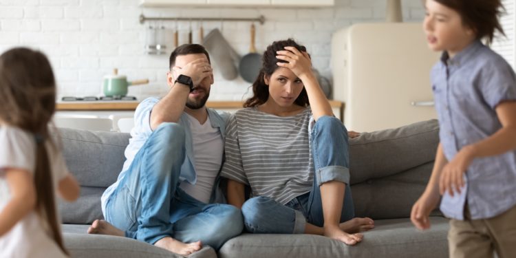 Genervte junge Eltern auf dem Sofa und zwei herumtollende Kinder