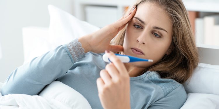 Im Bett liegende Frau blickt auf ein Fieberthermometer