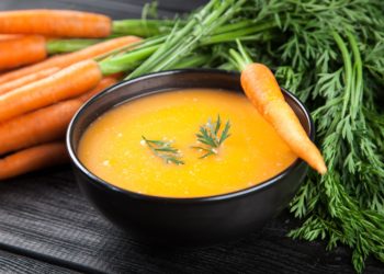 Eine schwarze Schale mit Möhrensuppe neben einem Bund frischer Möhren auf einem schwarzen Holztisch