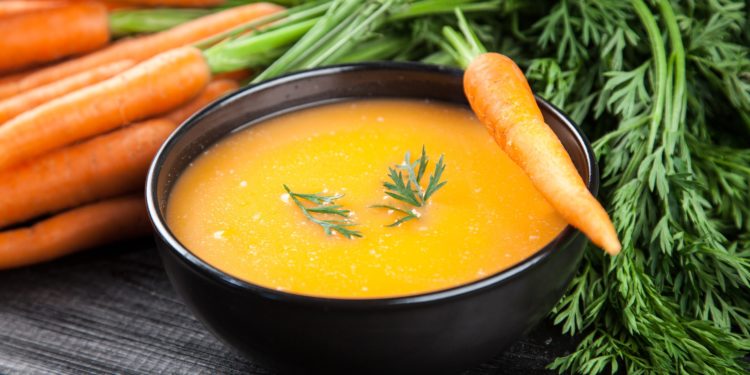 Eine schwarze Schale mit Möhrensuppe neben einem Bund frischer Möhren auf einem schwarzen Holztisch