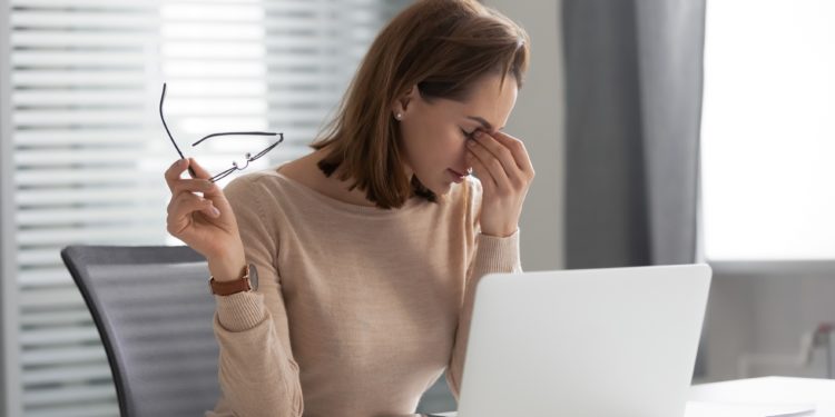 Erschöpfte junge Frau sitzt vor ihrem Laptop und fasst sich an ihre Nasenwurzel