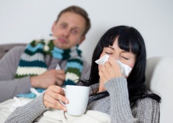 Mann mit Schal und Frau mit Teetasse und Taschentuch liegen zusammen auf dem Sofa