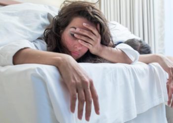 Eine Frau liegt im Bett und hält sich die Hand vor das Gesicht.