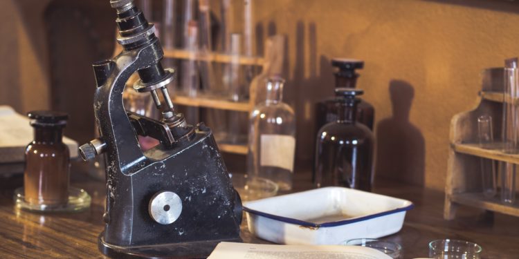 Historisches Labor mit Mikroskop, Reagenzgläsern, braunen Apothekerflschen, aufgeschlagenem Buch auf einem Holztisch.