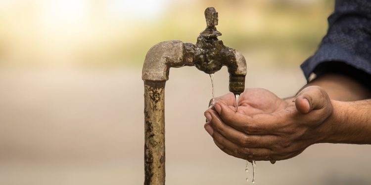 Eine Person hält die Hände zu einer Schale geformt unter einen verrosteten Wasserhahn, um etwas Wasser aufzufangen.