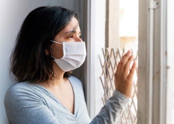 June Frau mit Mund-Nasen-Schutz sieht aus dem Fenster