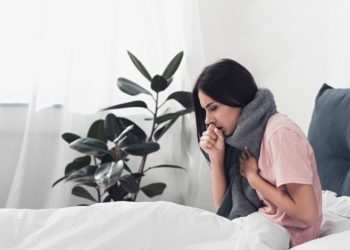 Hustende Frau mit Halstuch sitzt im Bett