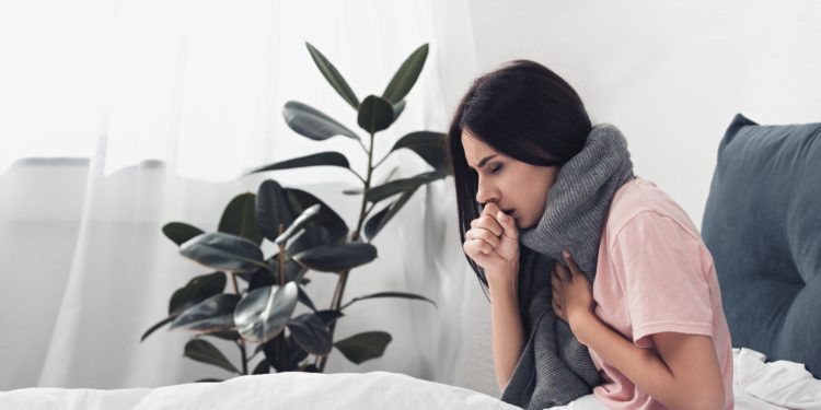 Hustende Frau mit Halstuch sitzt im Bett