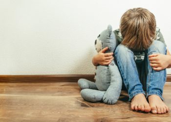 Kleiner Junge sitzt auf dem Boden, mit dem Kopf auf den Knien, hält einen Teddy im Arm