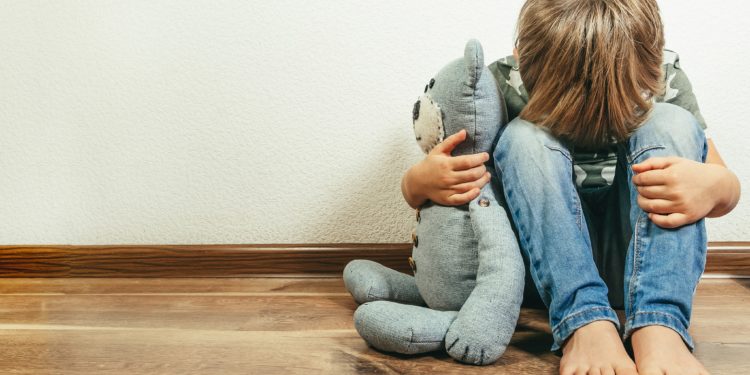 Kleiner Junge sitzt auf dem Boden, mit dem Kopf auf den Knien, hält einen Teddy im Arm